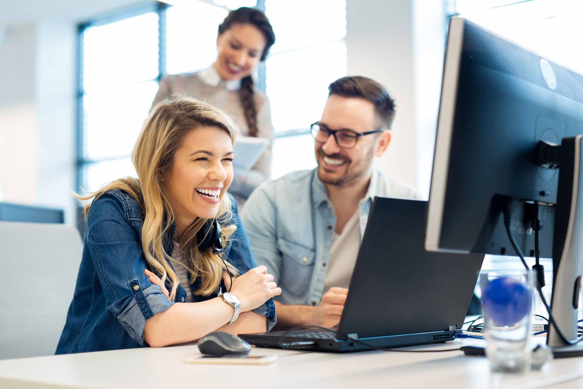 Drei fröhliche Kollegen arbeiten zusammen an einem Laptop und einem Monitor in einem hellen Büro, ein Symbol für Teamarbeit und positive Arbeitsatmosphäre.