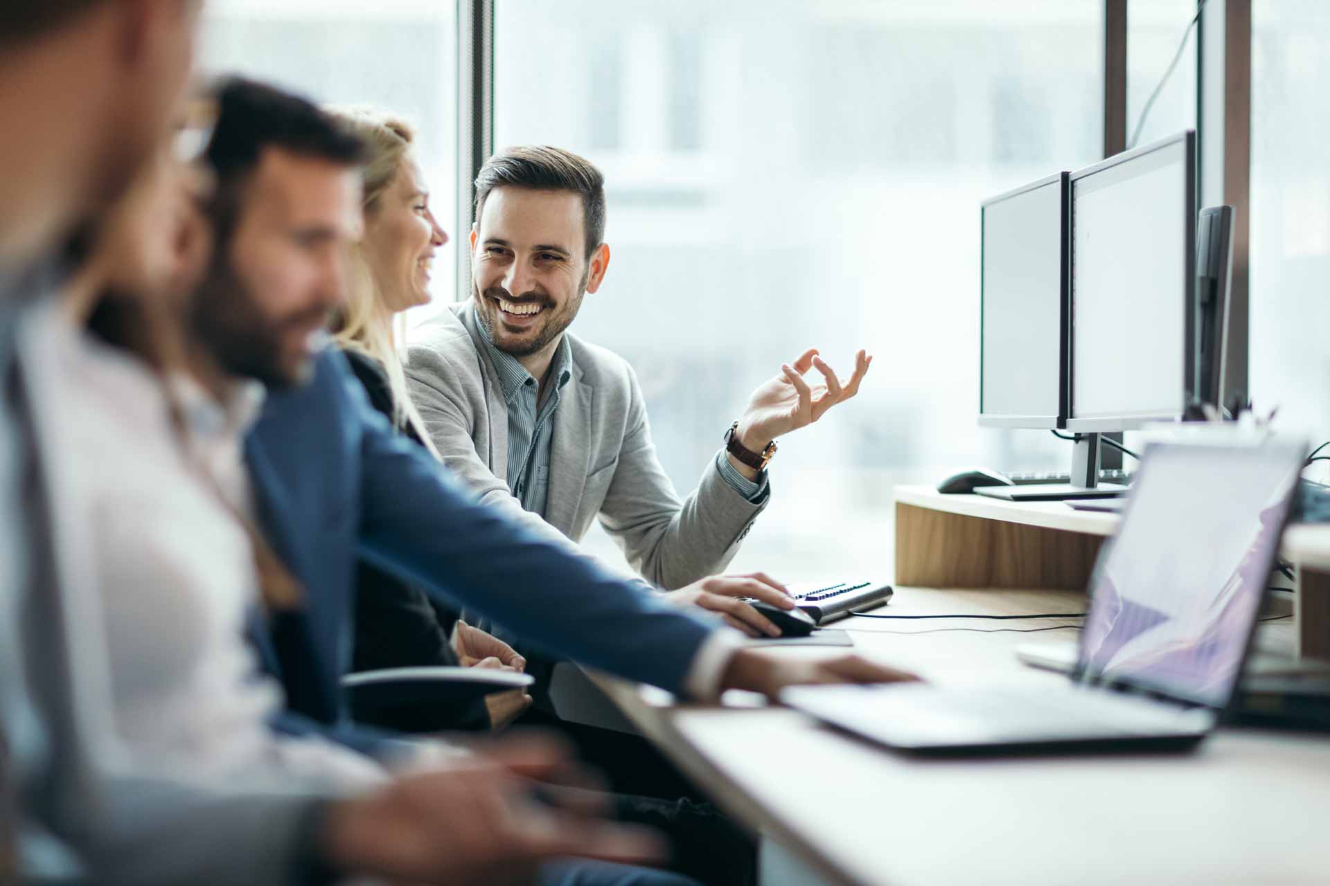 Büro-Team an Laptop und Monitoren, Mann führt Diskussion mit Geste.
