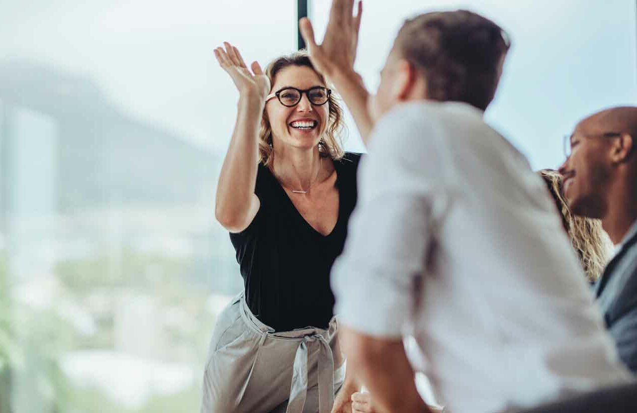 Eine fröhliche Frau gibt Kollegen High-Five, umgeben von Kollegen in einem hellen Büro.